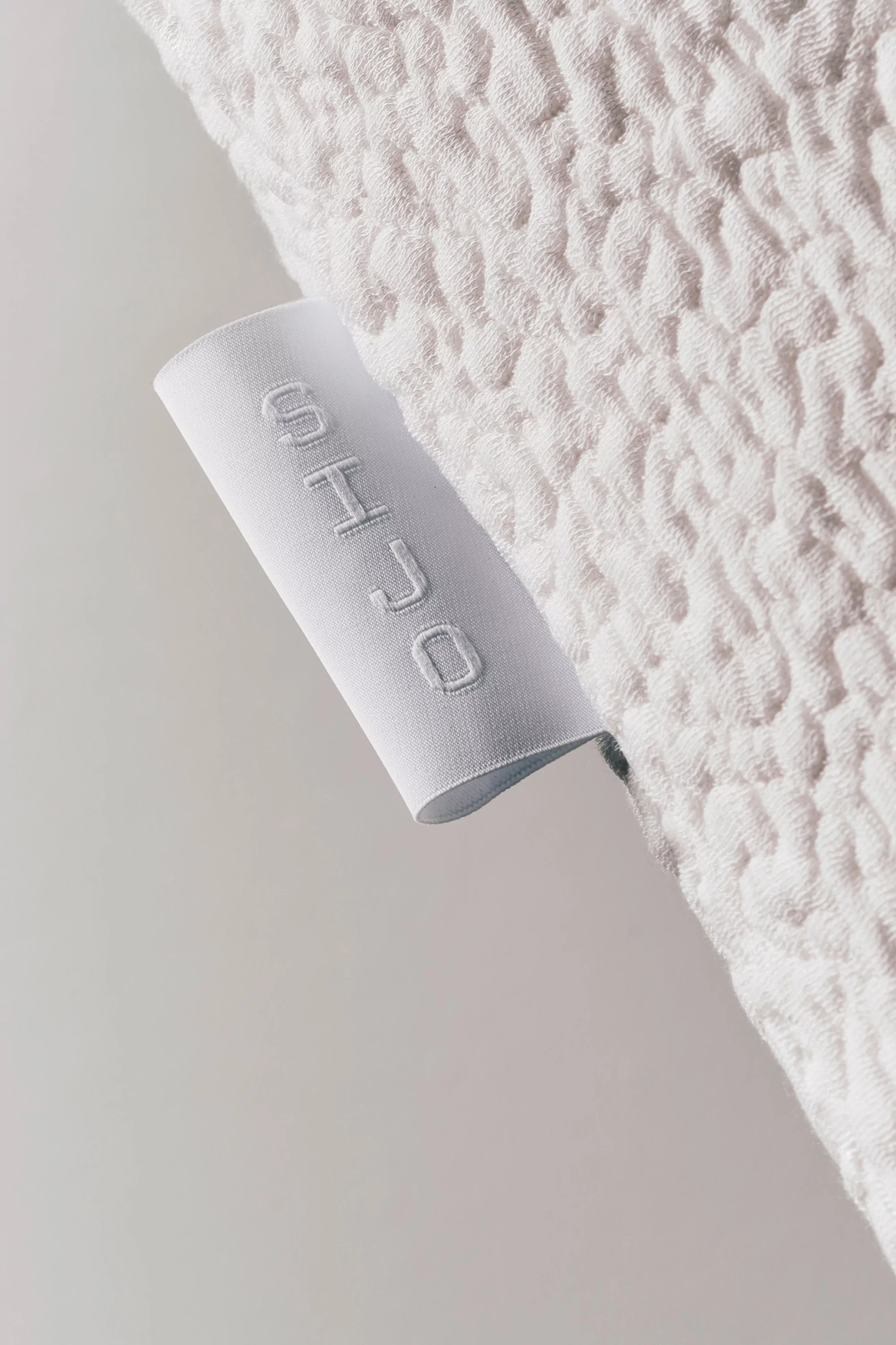Close-up of a white fabric with a textured pattern and a tag with the word 'SHJO' stitched vertically on a neutral background.
