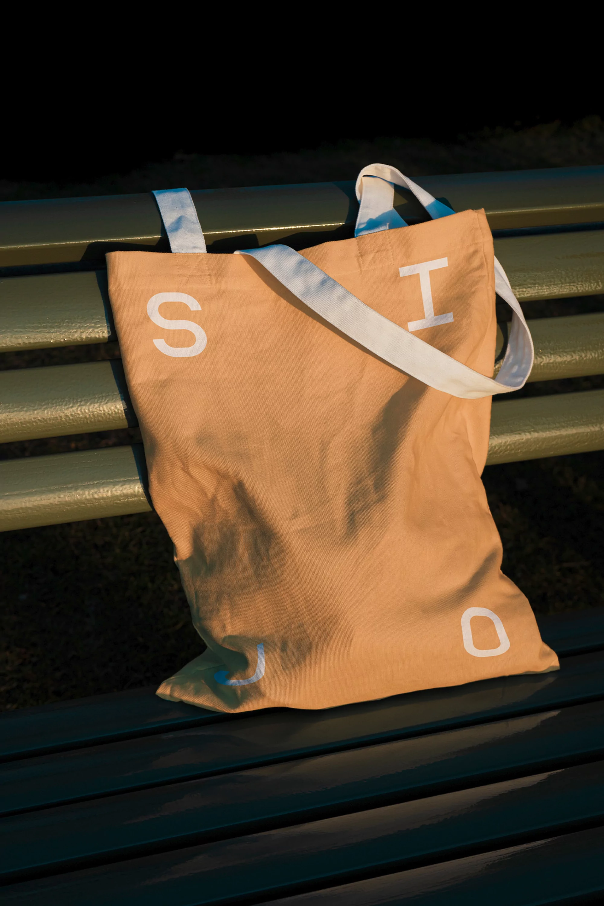 Tan tote bag with white lettering and handles on a green park bench in dappled sunlight, creating a simple, outdoor scene.