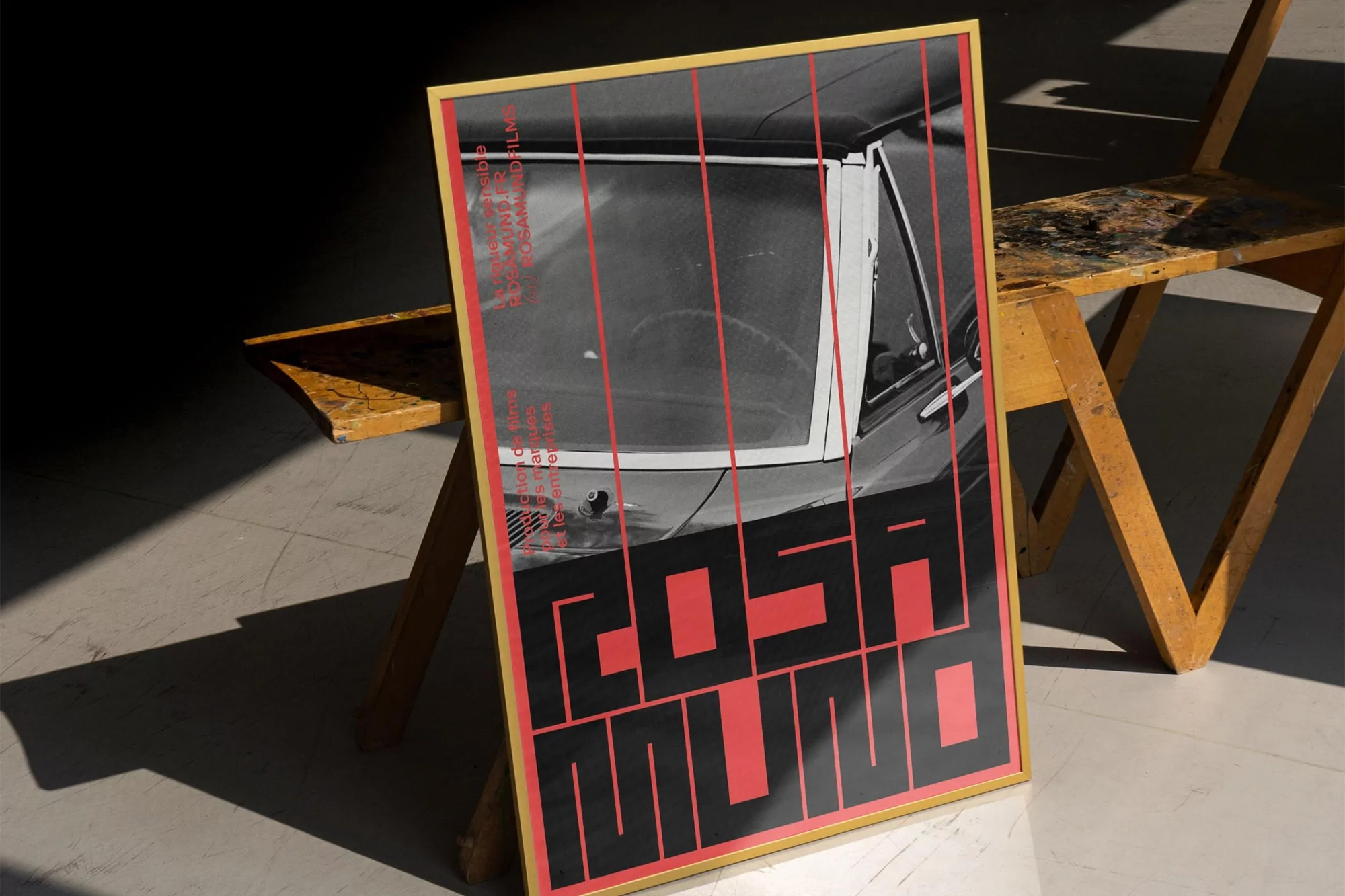 Framed movie poster features a vintage car image, bold red text, and sits beside a wooden folding chair in a studio with shadows.