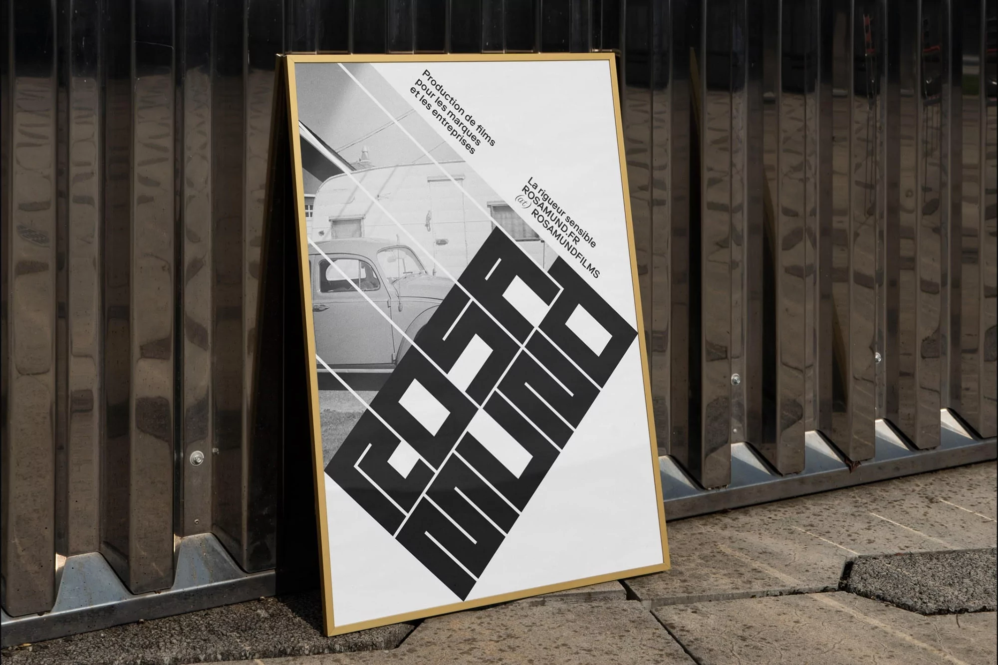 Framed vintage advertisement poster with gold trim leans against metallic wall. The poster features an image of a car and production company text.