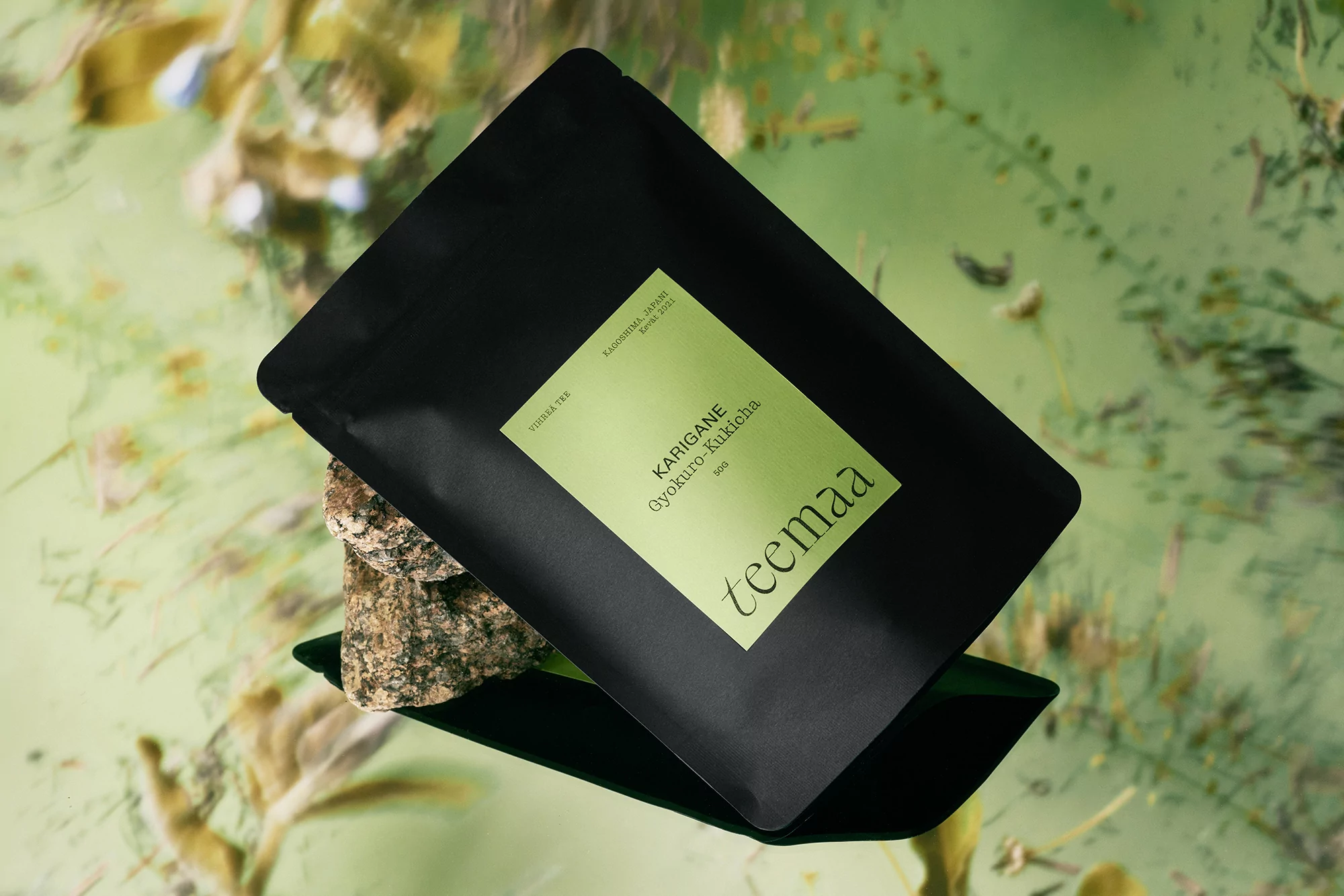 A black sealed tea bag with a green label lies on a rough stone, against a light green foliage background, creating an inviting, natural scene.
