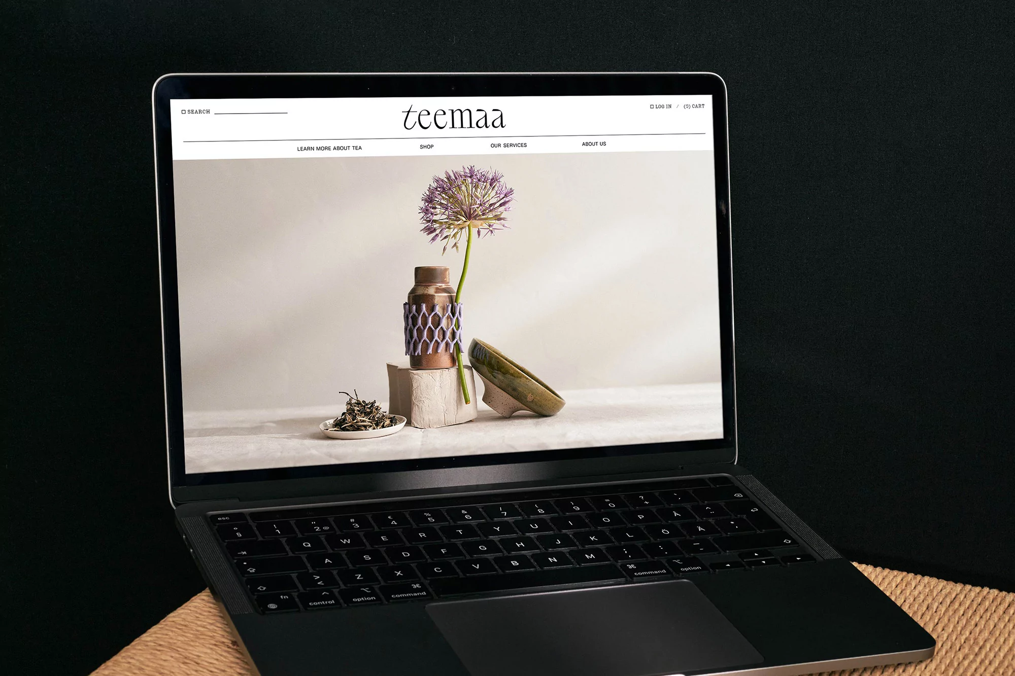 Laptop displays the Teemaa website with a tea product display, styled with a purple flower, against a black background.
