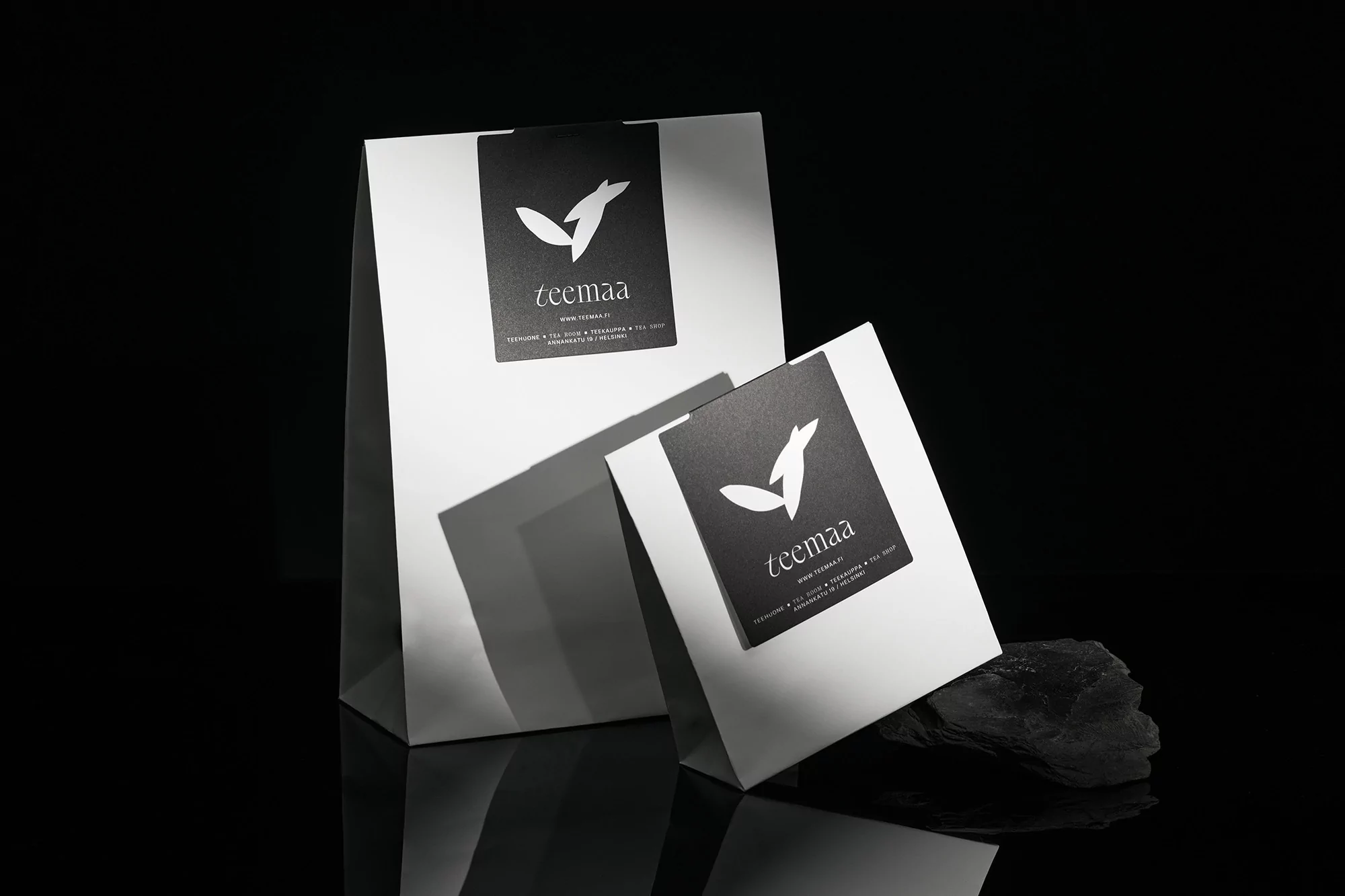 Two white paper shopping bags with black labels displaying the 'teemaa' logo, set against a dark reflective surface and background.