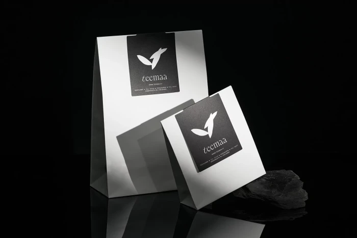 Two branded white paper bags with black labels set against a dark reflective background with a dark stone piece.