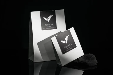Two branded white paper bags with black labels set against a dark reflective background with a dark stone piece.