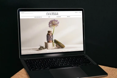 A laptop displaying the TeaMaa e-commerce website showing a product arrangement of tea packaging, a flower, and a ceramic bowl.