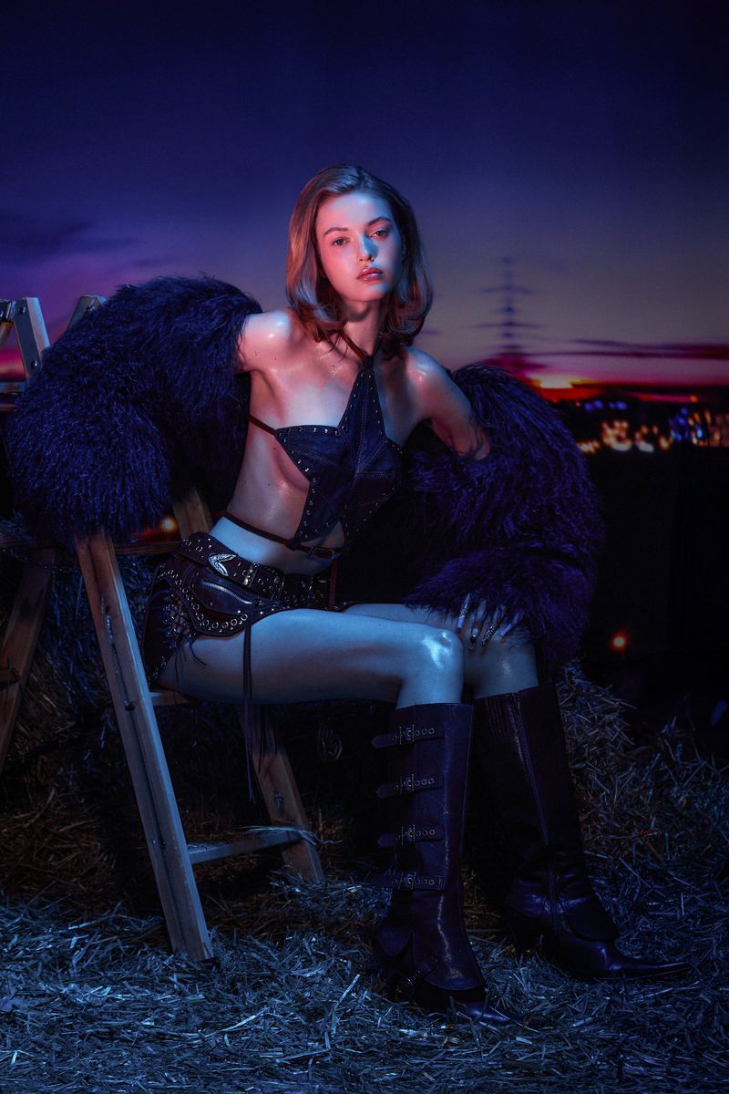 Fashionable young woman poses in avant-garde clothing, seated on a ladder in a dimly lit, rural setting at dusk.