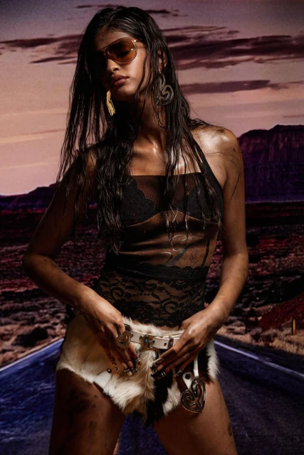 Woman poses in desert landscape with dramatic sky wearing sunglasses, lace top, fur shorts, belt and earrings.