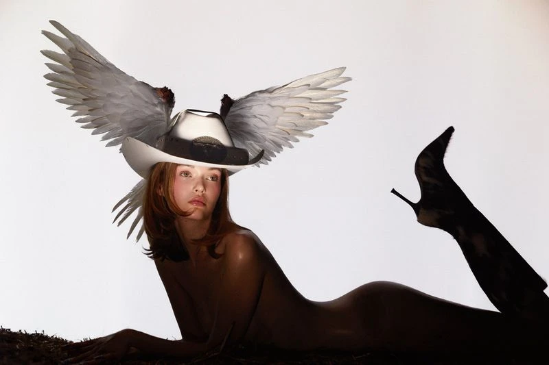 Woman poses reclining wearing a cowboy hat adorned with white wings, her figure accentuated by black high-heeled boots against a white backdrop.