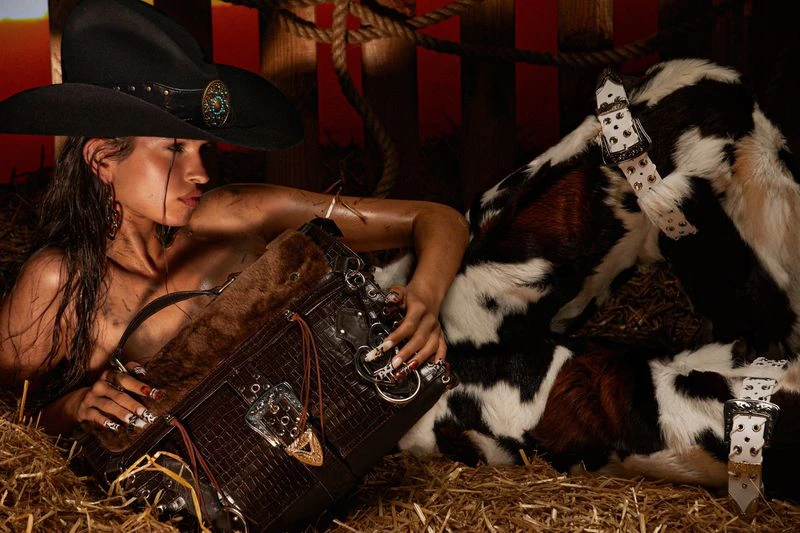 Adult female model with western hat poses with a brown purse and a goat against a rustic rope and wood background in a studio.