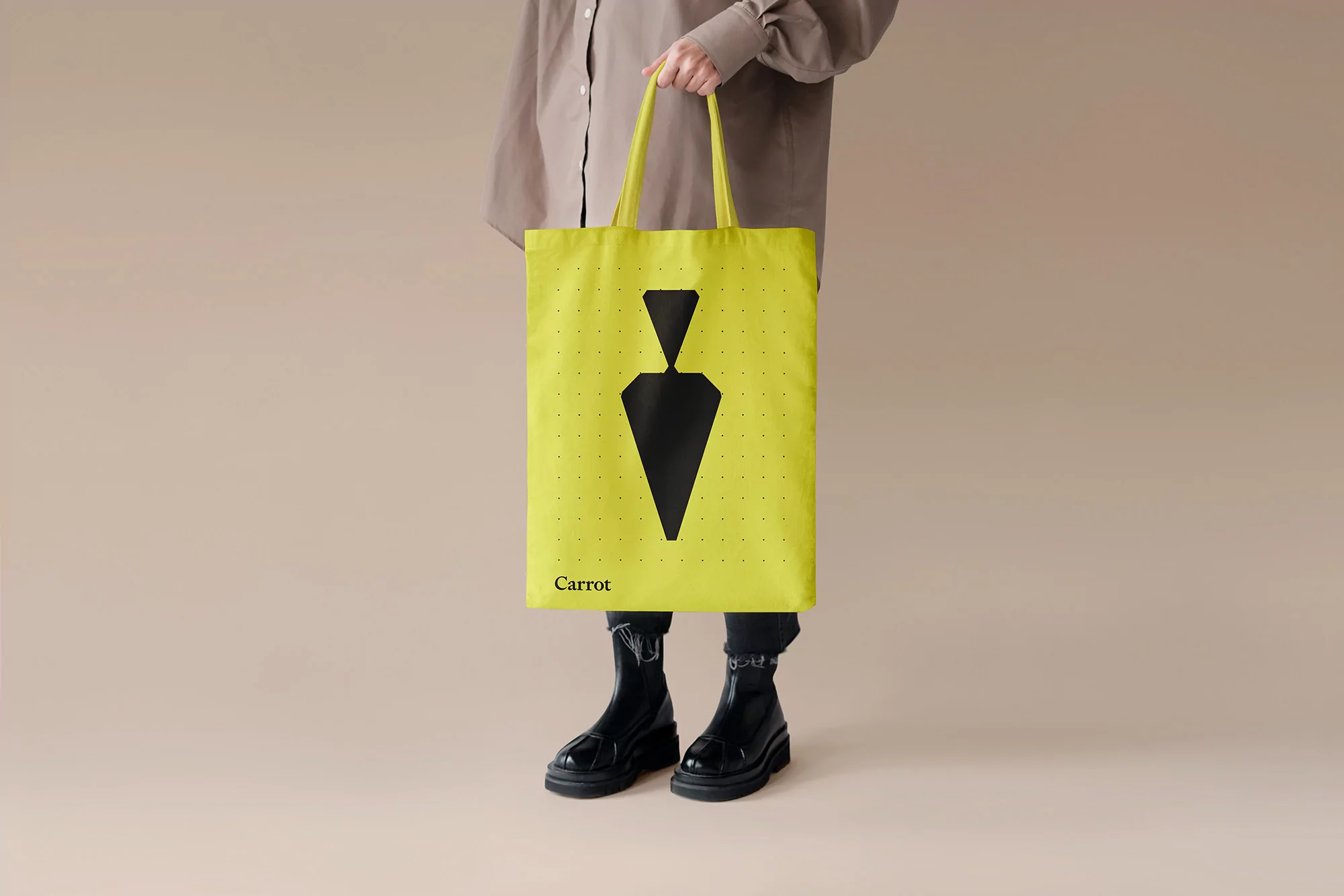Person holds yellow tote bag with black carrot design, paired with a tan shirt and black boots, set against a neutral background.