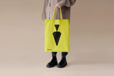 Person holding a bright yellow tote bag with a black geometric logo against a plain neutral studio background.
