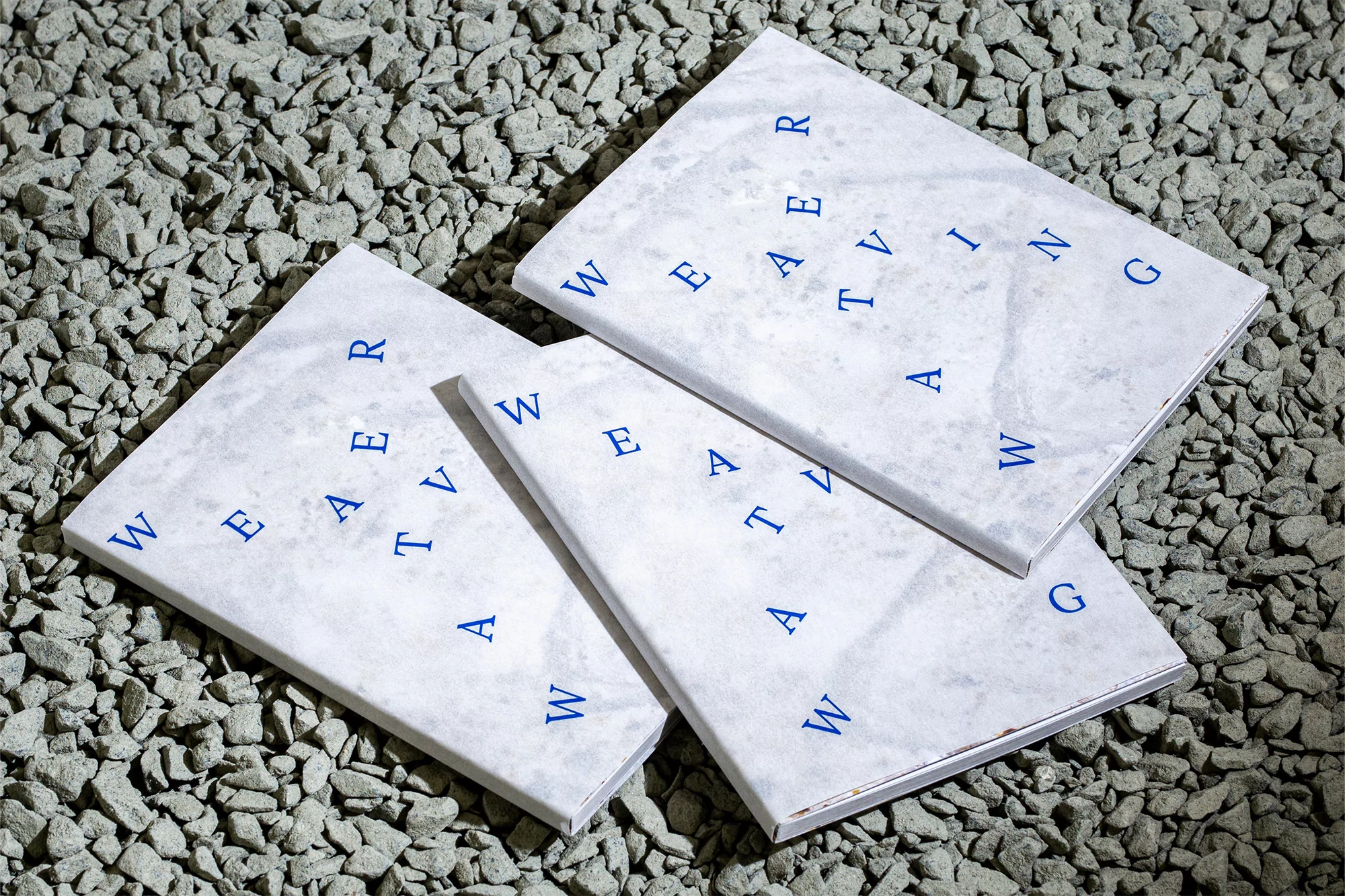 Three square books with blue lettering spelling “Weaving” are arranged on a surface covered with small grey gravel.