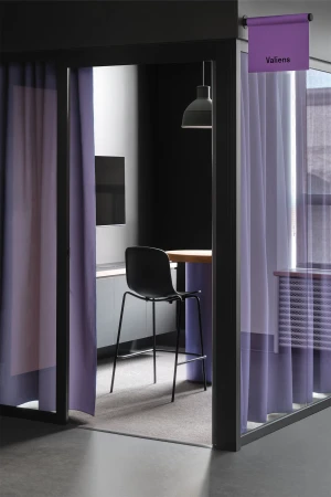 Interior view of a booth-like office space with a chair at a wooden table, separated by mauve curtains, and a Valens sign.