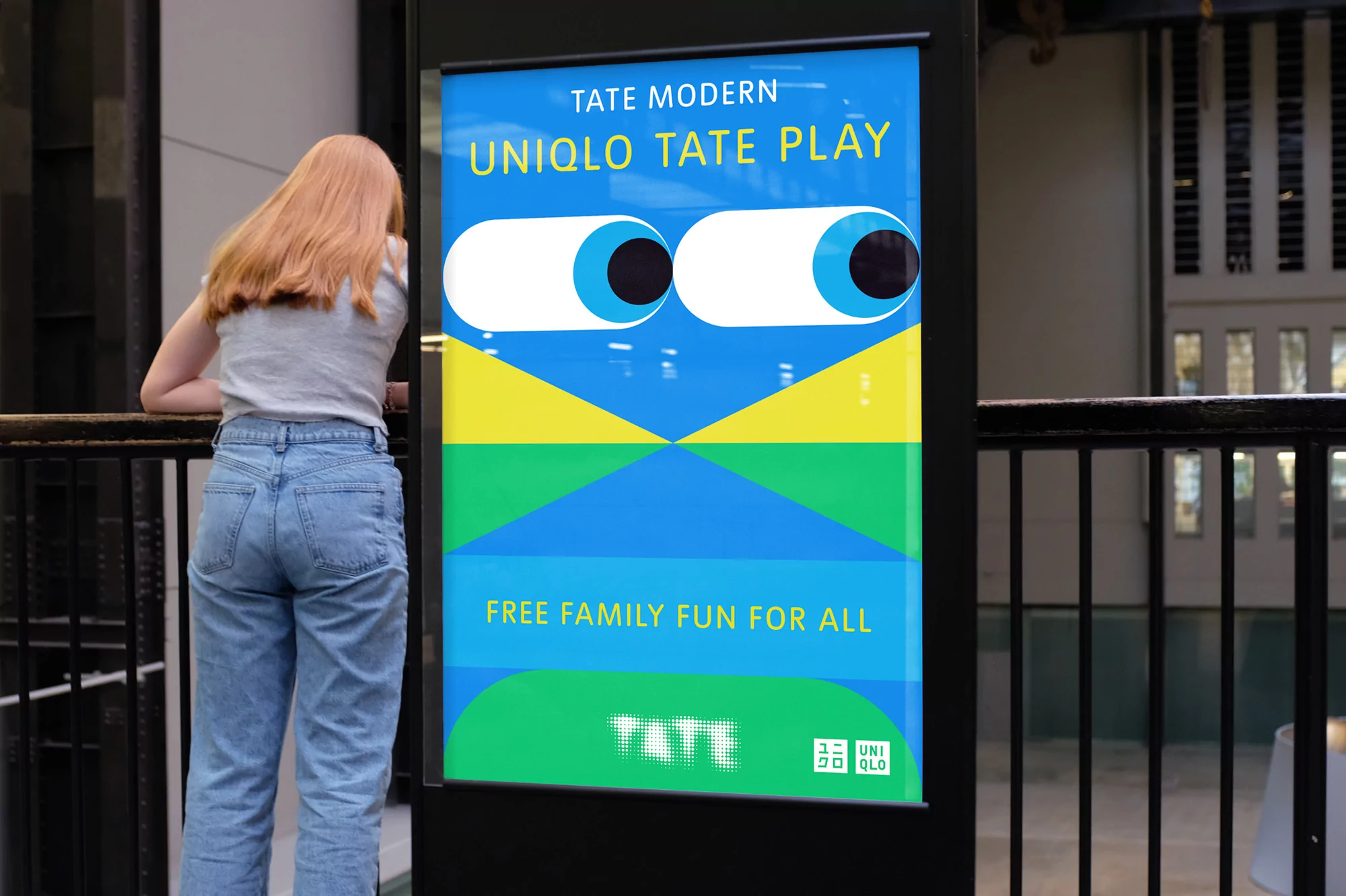 A woman stands near a Uniqlo Tate Play advertising sign for the Tate Modern, featuring cartoon eyes and geometric shapes in blue, green, and yellow.