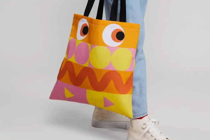 Person carrying an orange tote bag with abstract geometric designs and black straps, paired with light wash jeans and high-top sneakers.