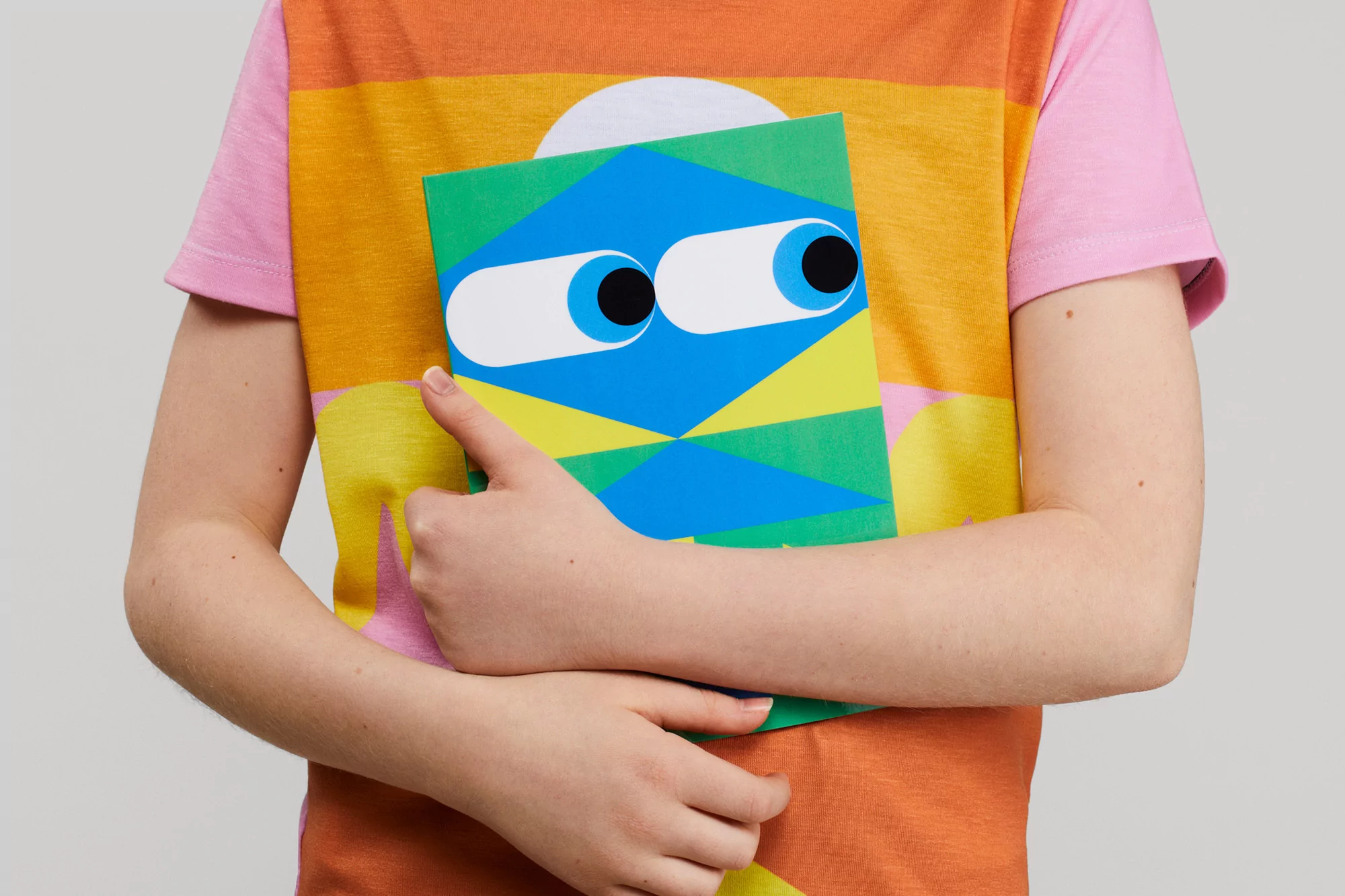 Young child holds a green book with an animated face in front of their brightly coloured t-shirt, set against a plain background.