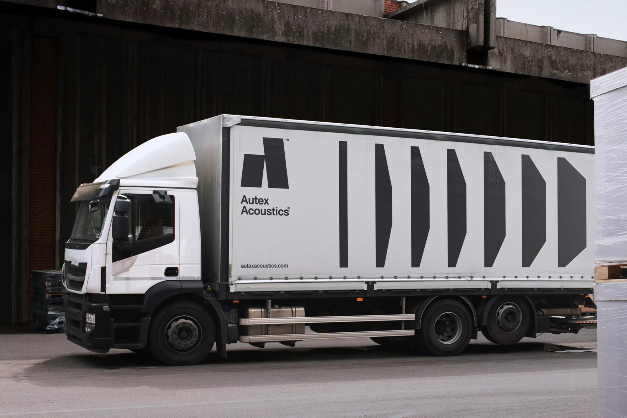 White commercial truck with black logo and stripes parked in an industrial area near a warehouse, transporting materials.