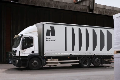 White Autex Acoustics branded delivery truck parked on an asphalt lot in front of a concrete building structure.
