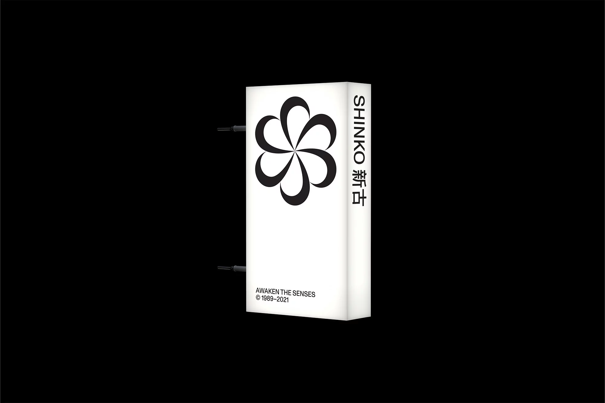 Illuminated vertical white sign with black flower icon, company name SHINKO and slogan AWAKEN THE SENSES in black against black background.