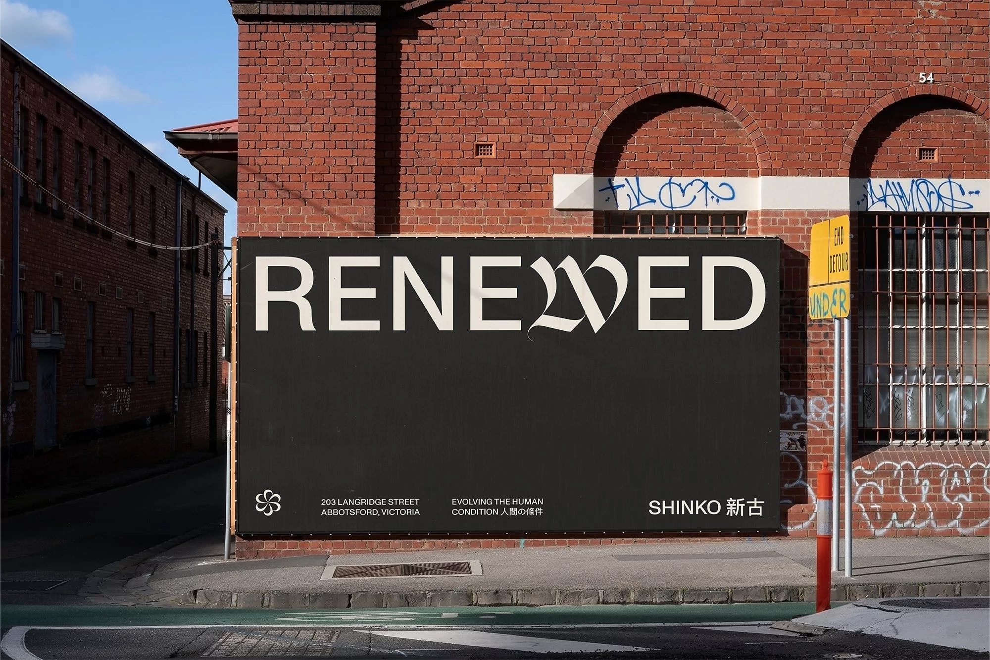 Urban black billboard featuring the word 'Renewed' in stylized white font, along with brand information and address details on brick building backdrop.