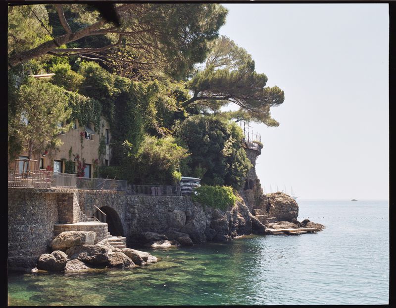 Coastal view of a stone building covered in greenery, steps leading down to the turquoise water, and a boat on the horizon.