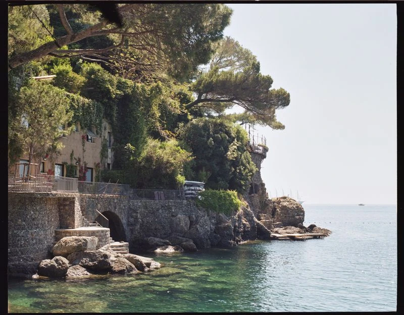 Coastal view of a stone building covered in greenery, steps leading down to the turquoise water, and a boat on the horizon.