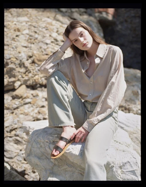 Young woman poses while sitting on a rock formation in sandals, light coloured shirt and pants, looking at the camera in natural light.