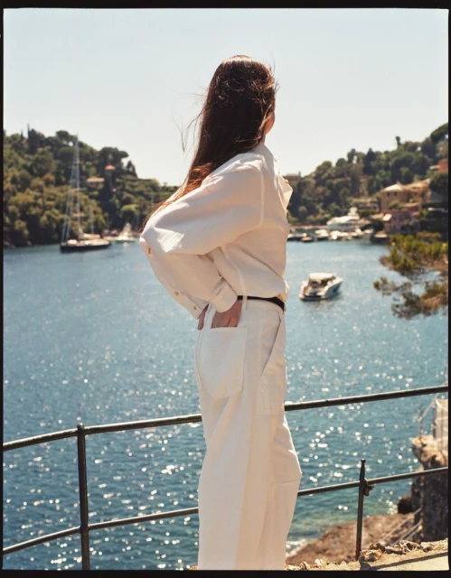 A woman in cream-colored pants and jacket faces the sea in a full outdoor shot, boats visible on the water.