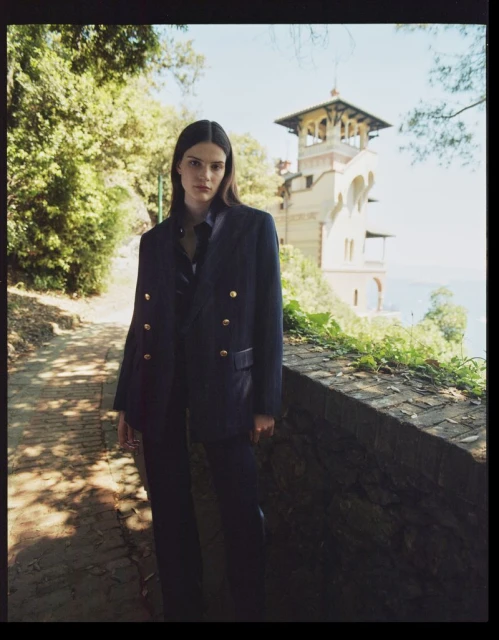 A fashion shot of a young adult female model in a navy suit posing on a stone pathway lined with greenery, with a villa in the background.