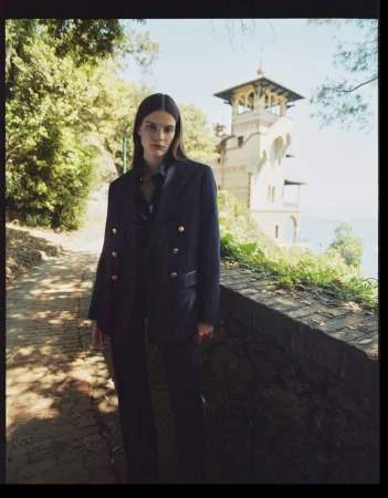 A fashion shot of a young adult female model in a navy suit posing on a stone pathway lined with greenery, with a villa in the background.
