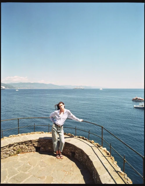 Adult female posing on stone platform near ocean under clear blue sky in outdoor scene with boats and distant land.