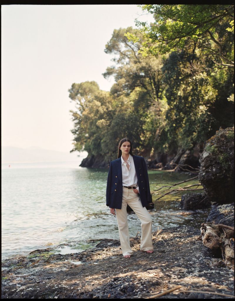 A woman in a blazer and white pants poses on a rocky lakeshore with a backdrop of lush trees and calm water under a clear sky.