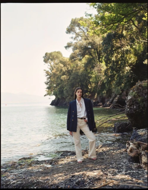 A woman in a blazer and white pants poses on a rocky lakeshore with a backdrop of lush trees and calm water under a clear sky.