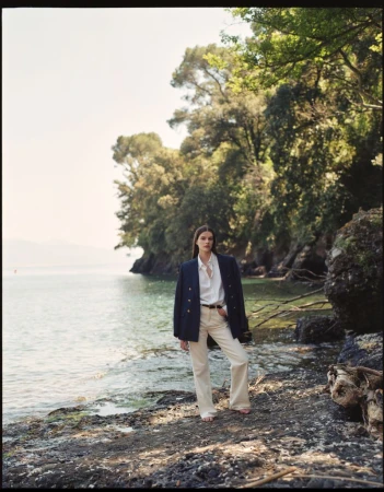 A woman in a blazer and white pants poses on a rocky lakeshore with a backdrop of lush trees and calm water under a clear sky.