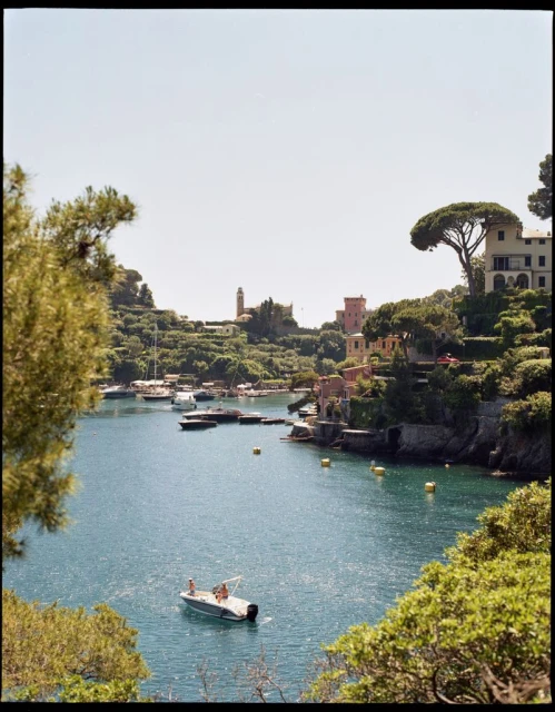 Picturesque Italian harbor view on a sunny day featuring a motorboat on calm water, surrounded by lush green trees and colorful buildings.