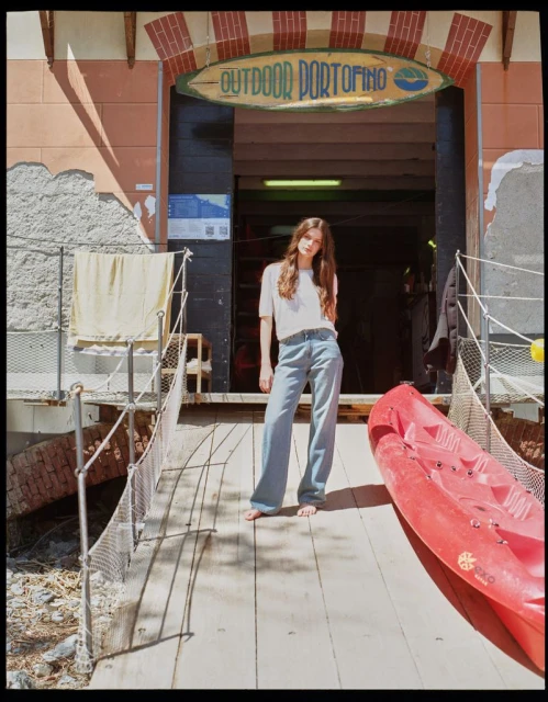 Adult woman stands barefoot on a wood deck next to a red kayak, in front of a store called Outdoor Portofino, on a sunny day.