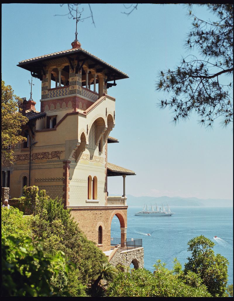 Scenic view of a historic building with an observation tower overlooking the blue sea, complemented by lush green trees and sailboats.