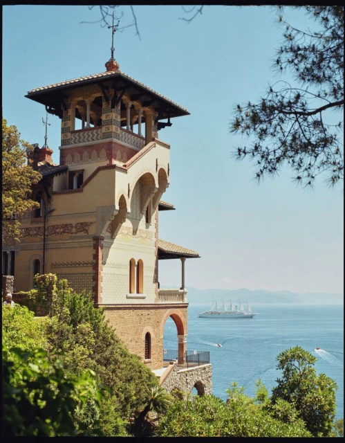 Scenic view of a historic building with an observation tower overlooking the blue sea, complemented by lush green trees and sailboats.