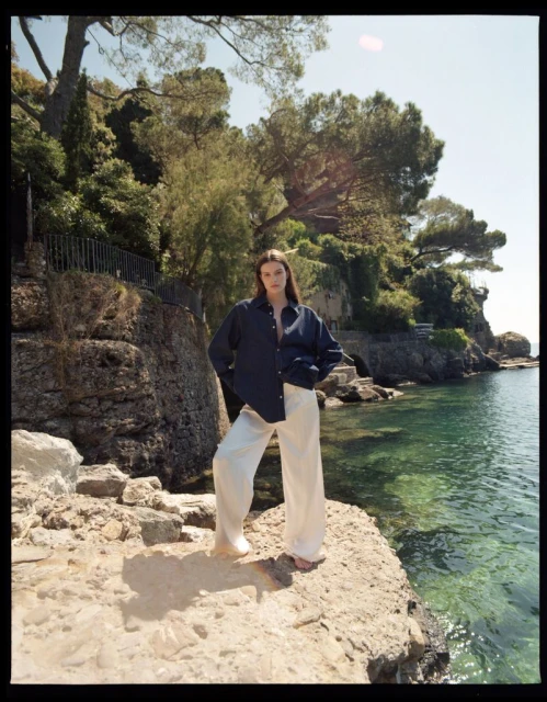 A young woman stands on a rocky outcrop wearing a navy button-down shirt and wide-leg cream trousers, with a coastal scene in the background.