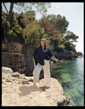 A young woman stands on a rocky outcrop wearing a navy button-down shirt and wide-leg cream trousers, with a coastal scene in the background.