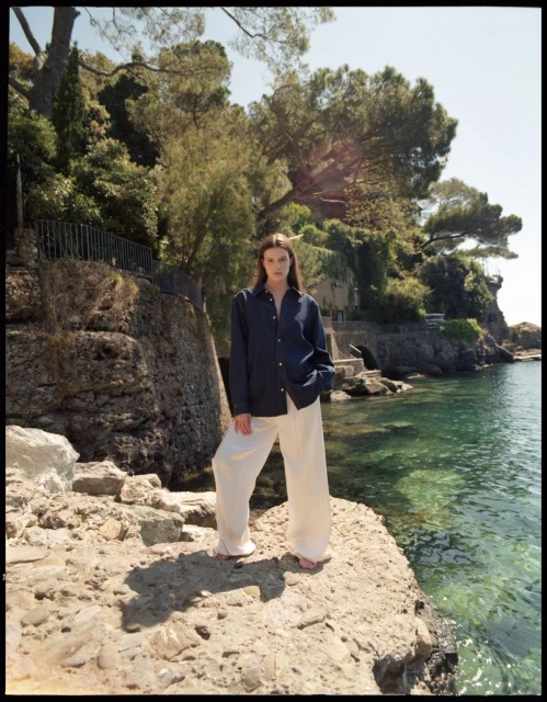 A woman in a navy shirt and white pants stands on a rocky outcrop overlooking the ocean, with lush greenery in the background.