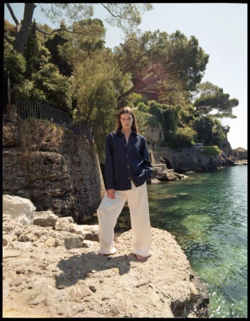 A woman in a navy shirt and white pants stands on a rocky outcrop overlooking the ocean, with lush greenery in the background.