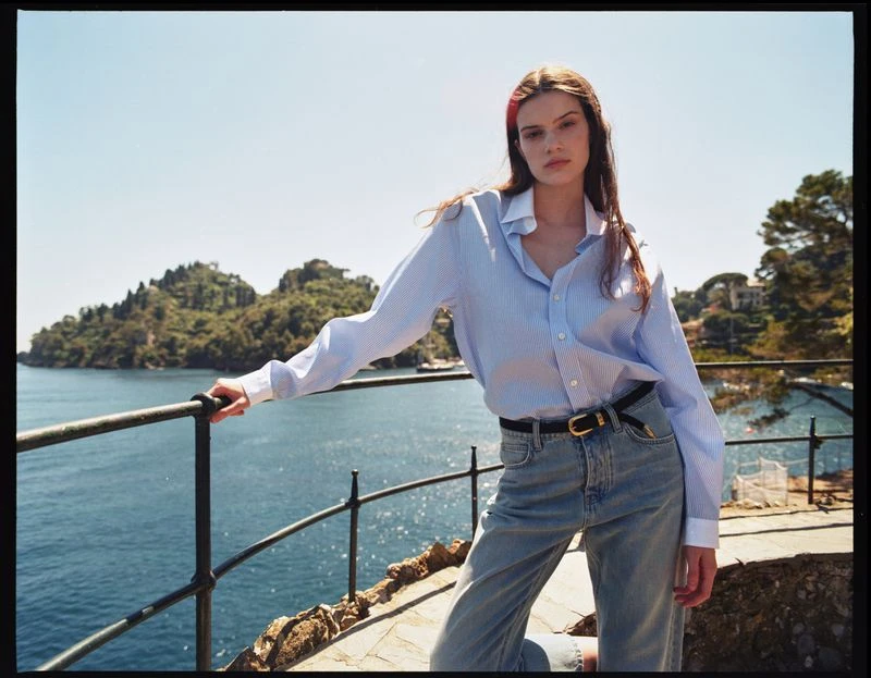 Fashion shot of a woman in a blue striped shirt and jeans leaning against a railing with an ocean and green landscape in the background.