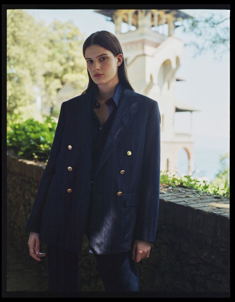 A woman in a double-breasted blazer stands outdoors in front of a stone wall and building with arches, showcasing a fashion-editorial aesthetic.