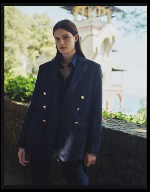 A woman in a double-breasted blazer stands outdoors in front of a stone wall and building with arches, showcasing a fashion-editorial aesthetic.