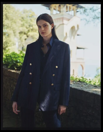 A woman in a double-breasted blazer stands outdoors in front of a stone wall and building with arches, showcasing a fashion-editorial aesthetic.