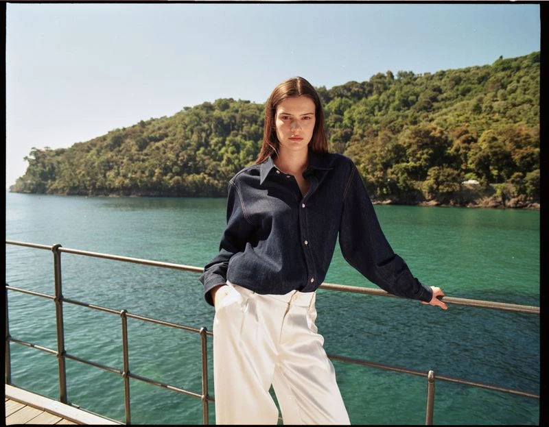A woman in a dark-blue, button-down shirt and white pants stands near a metal railing with a forested coastline and turquoise sea in the background.