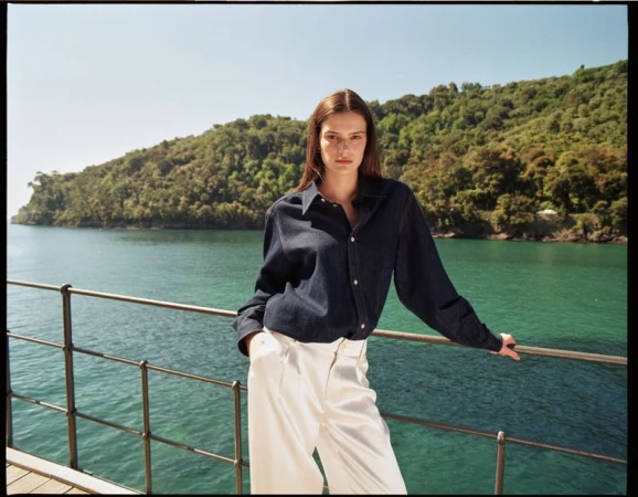 A woman in a dark-blue, button-down shirt and white pants stands near a metal railing with a forested coastline and turquoise sea in the background.