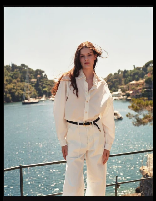 A woman stands in a bright, outdoor fashion shot, wearing a white blouse and trousers against a harbor and sky backdrop.